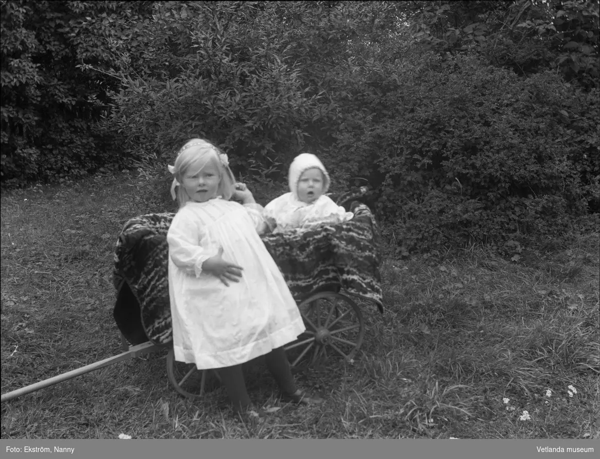 Två barn och en barnvagn i en trädgård. Barnens anhöriga var troligtvis bekanta med fotografen.