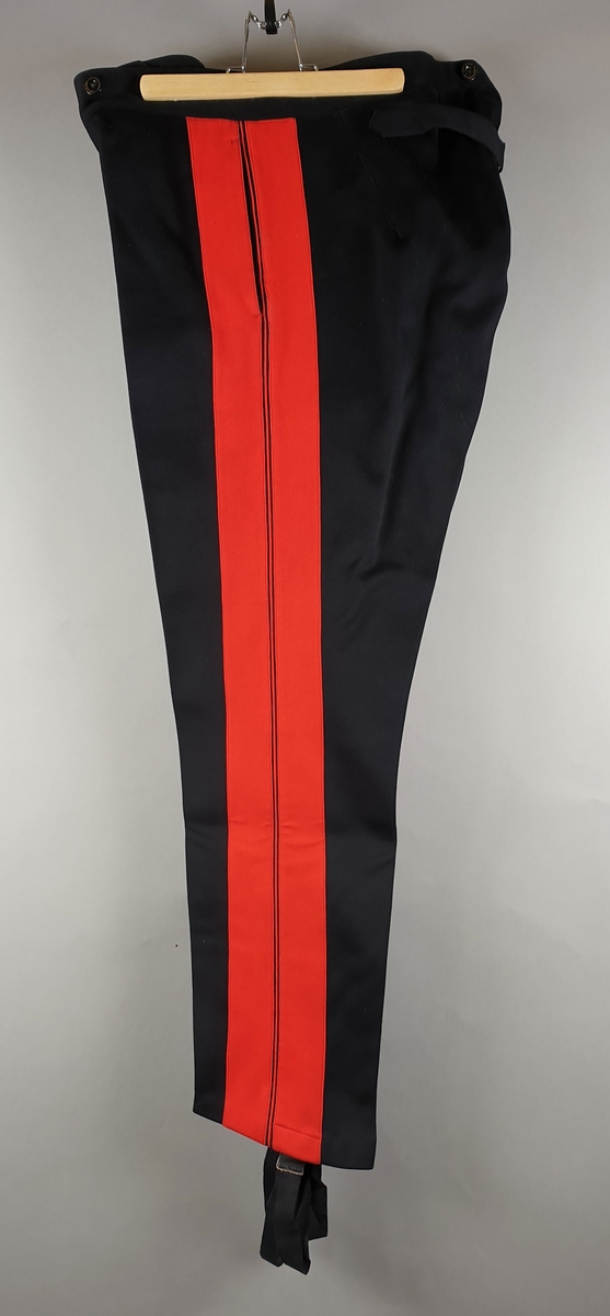 Uniformsbukse med brede røde striper på hver ytterside. Buksen smalner inn langs leggene og har knapper nederst. Knapper i gylfen og innsydde lommer.