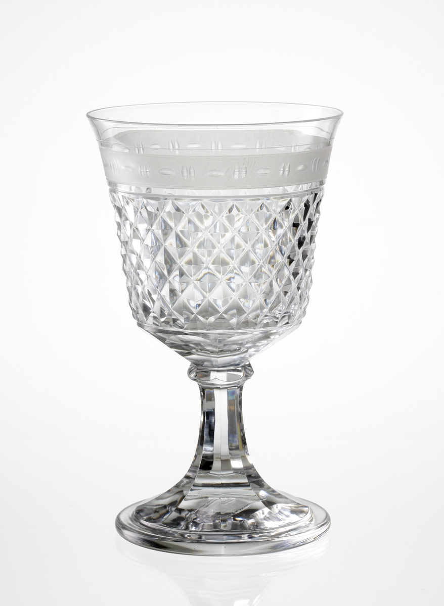 Design: Vicke Lindstrand, Edward Hald. 
Rödvinsglas, klockformad kupa med diamantslipning, graverad bård och slipade fasetter runt botten. 
Fasettslipat ben med knapp.