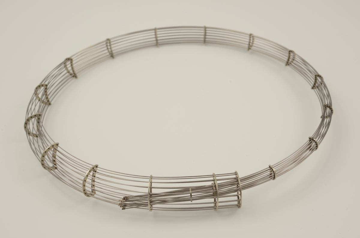 Halssmykke laget av 16 tynne metalltråder som er tredd gjennom 19 halvmåneformede metallstykker. Metallstykkene minsker i størrelse, slik at smykket smalner i den ene enden. Smykket danner en ring ved at den smale enden er tredd inn i den brede.