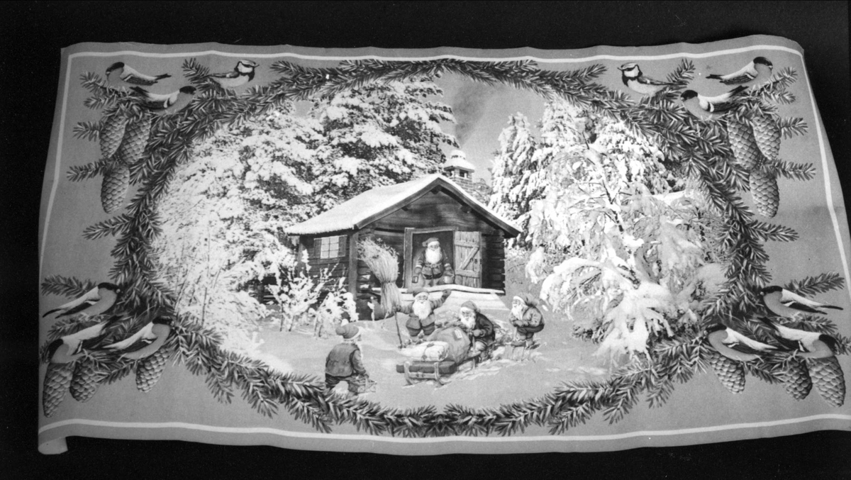 Fem tomtar i vinterlandskap. En tomte står i dörröppningen till en stuga. Övriga är sysselsatta med en julklappsssläde, fågelkärve, snöskottning m.m. Bilden inramad av granris i vilket det sitter domherrar och blåmesar.