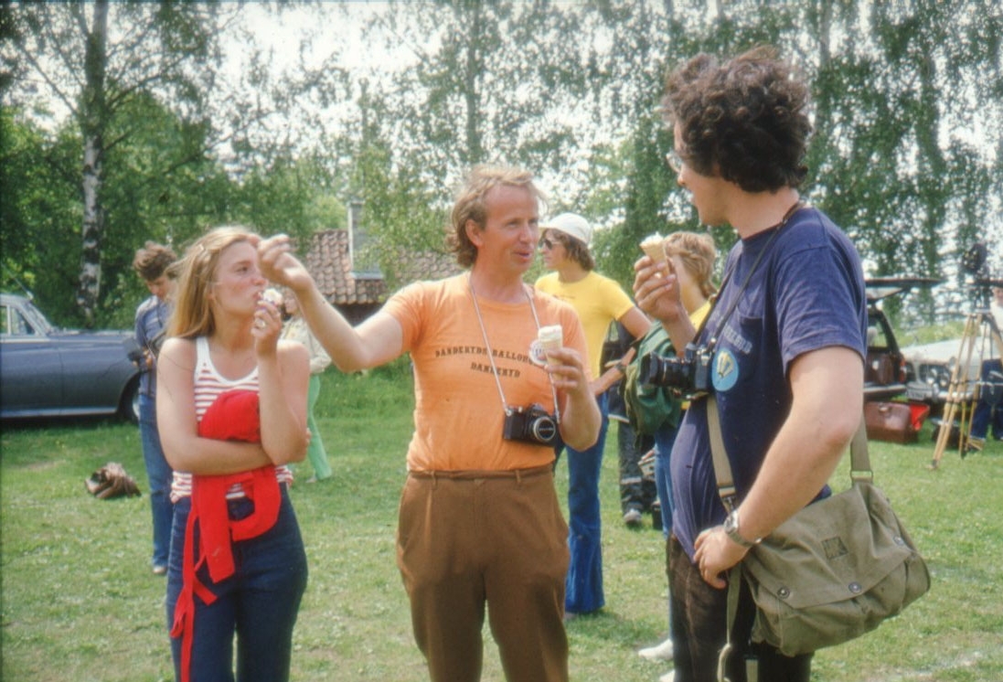 En kvinna och två män pratar och gestikulerar, de äter strutglas. Männen har kamera runt halsen och mannen i mitten har en svagt orangea t-shirt med texten, troligen: "Danderyds Ballongklubb". Det är sommar och sol. Diabild.