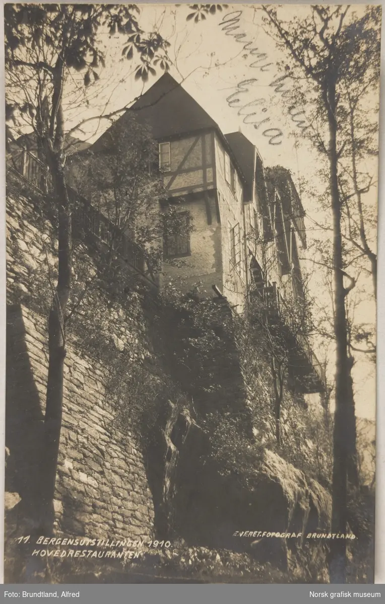 Postkort med bilde av et hus i bindingsverk som ligger ytterst oppå en steinmur.