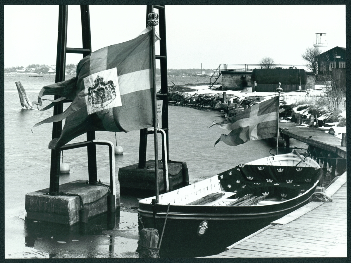 Bilden visar en valbåt som har utrustats som kungaslup vid kajen framför i Slup- och Barkasskjulet på Stumholmen i Karlskrona. Örlogsflaggan med riksvapnet är utvecklad på akter. Vid bogen finns en svensk flagga till. Sittbänken i båten är belagt med tyg som är mönstrad med kronor. Årorna ligger i båten.