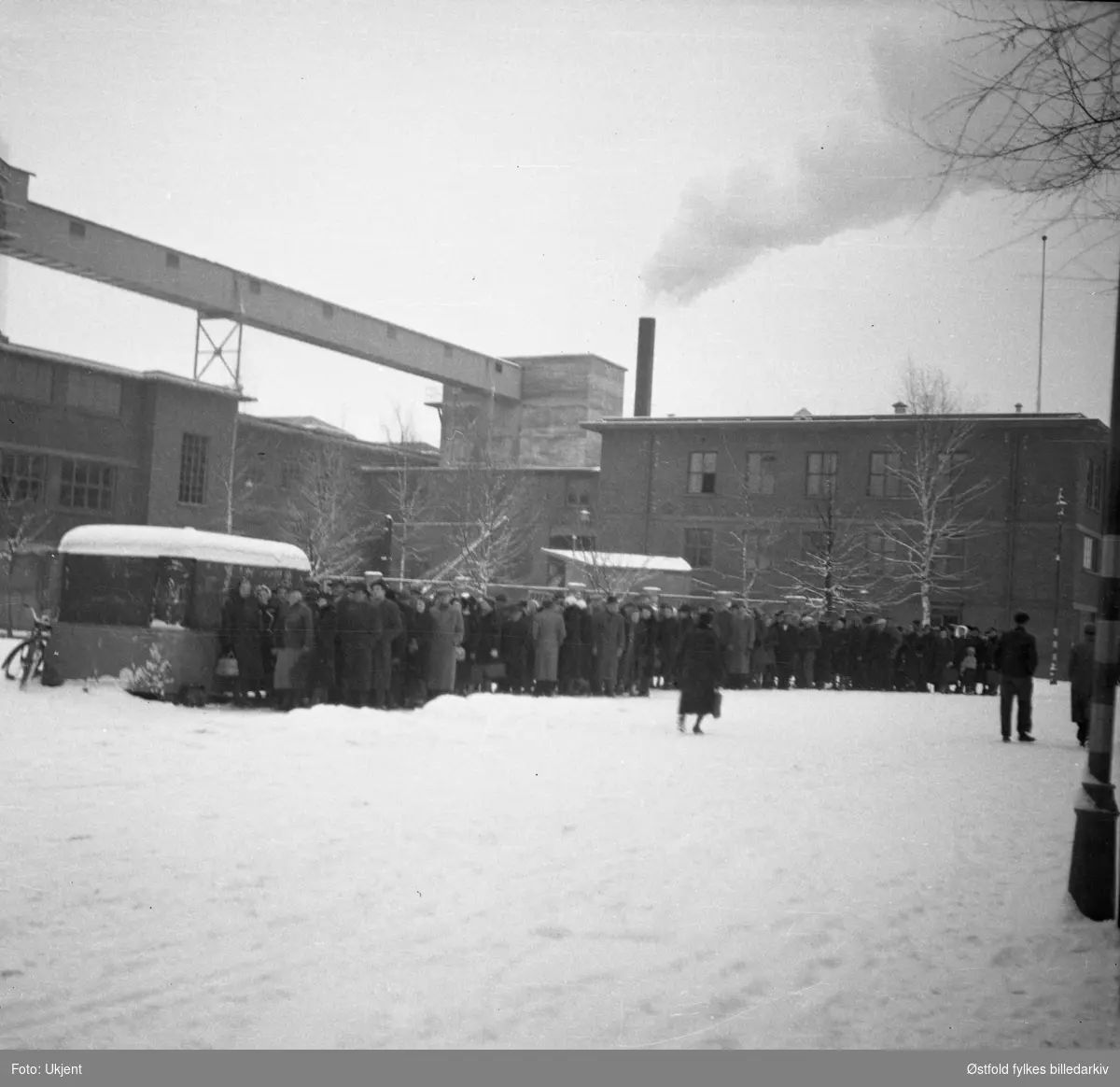 Folk i kø for kjøtt og fisk under 2. verdenskrig i Askim 1940-1945. Askim gummivarefabrikk i bakgrunnen.