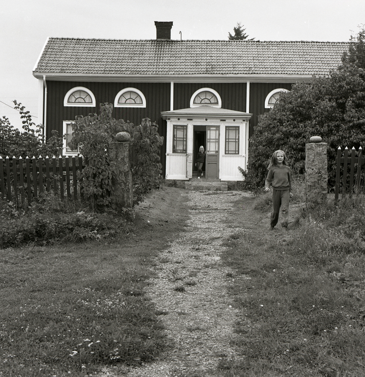 Boningshus med inbyggd förstukvist samt halvmånefönster på övervåningen, Järvsö 1980. En flicka står i husets dörröppning och en annan kommer springande genom grindhålet.