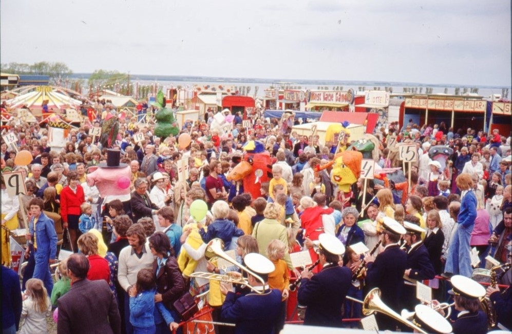 Ett tivoliområde med folksamling, papier-mache figurer och musikkår vid Vätterns strand. Fotograferat lite uppifrån, en scen troligen.