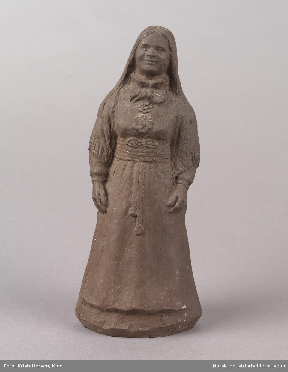 Figur av en kvinne med løst hår iført en bunad. Skulpturen er laget av leire. Bunaden ser ut som en beltestakk fra Telemark. Skulpturen er hul.