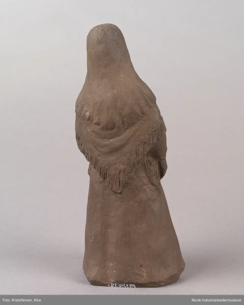 Figur av en kvinne med løst hår iført en bunad. Skulpturen er laget av leire. Bunaden ser ut som en beltestakk fra Telemark. Skulpturen er hul.