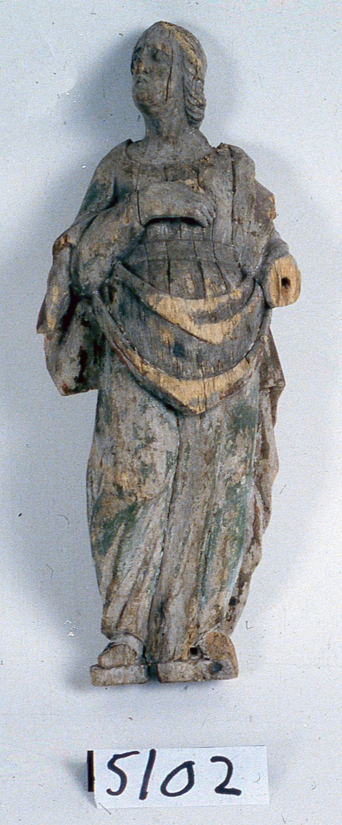 Skulptur av trä. Stående kvinnofigur i kontrapostställning med huvudet högervänt, blickar snett uppåt. Klädd i lång klänning med skärp. Manteln över vänster axel hålls samman med höger arm och faller i bågformiga veck framtill. Höger hand vilar på bröstet, vänster arm borta. Näsan avslagen, likaså främre delen av fötterna. Halva fotplattan borta. Många delar skadade. Spår av kredering, röd, brun, grön och svart färg. Har troligen tillhört epitafium eller altaruppsats.