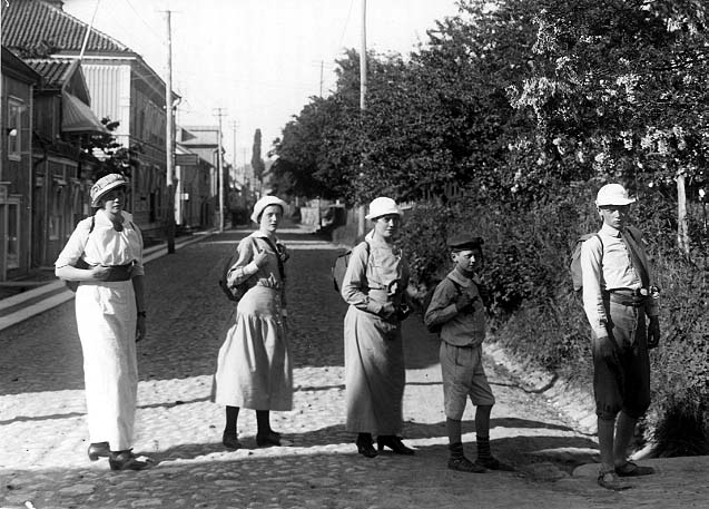 Tre flickor/ung kvinna och två pojkar med ryggsäckar står på rad över den kullerstensbelagda Brahegatan i Gränna. Eftermiddagssol från väster.