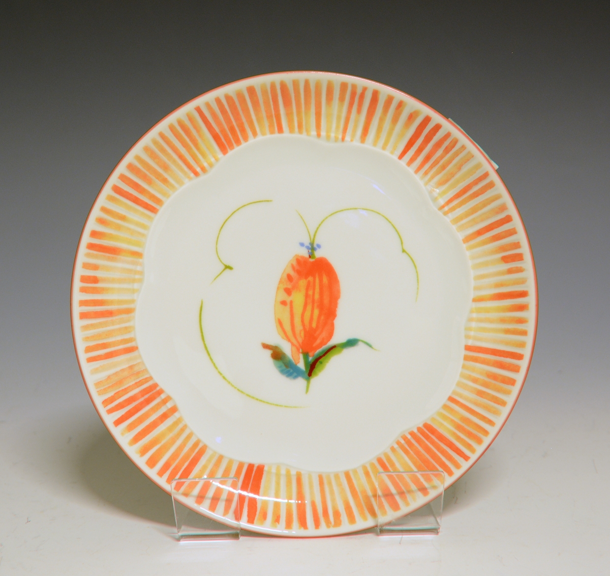 Frokosttallerken av porselen med hvit glasur. I senter en oransje blomst. På fanen oransje, radiære striper.
Modell: 2700 Fiore av Eeva Terävä
Dekor: Spring av Victoria Berge