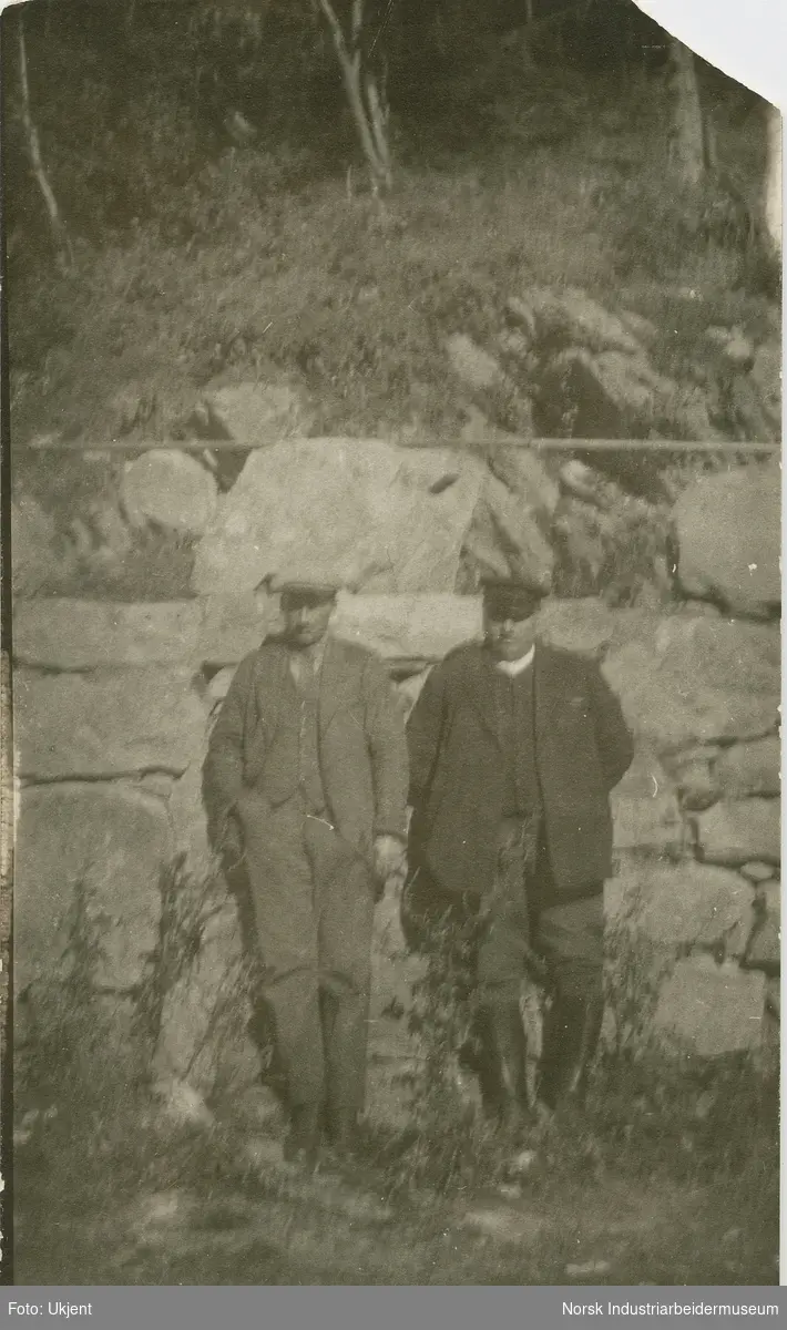 To menn i uniform stående foran steinmur.