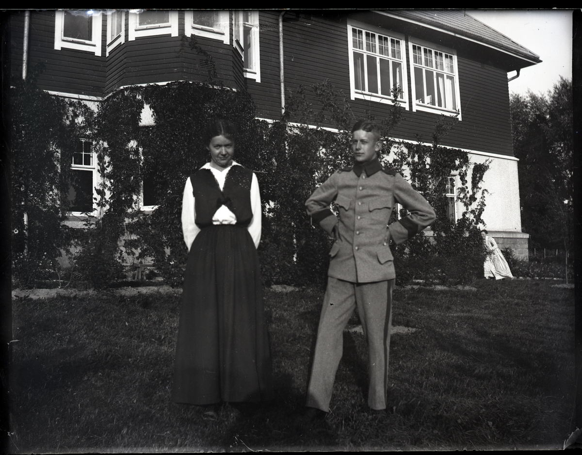 "Husmodersskolan".
Ung kvinna och man i uniform, stående utanför Husmodersskolan. Kvinna i randig klänning gömmer sig bakom buskage.