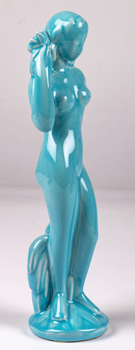Figurin Eva, stående flicka, höger hand håller en korg med blommor på axeln, vänster hand lutad mot stubbe/buske. Posen och attributen ofta vanlig för att symbolisera ymnighet. Turkos glasyr. Formgiven av Eva Staehr Nielsen.
