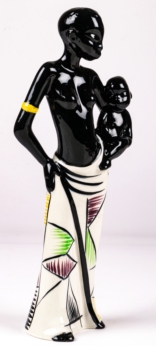 Figurin föreställande svart kvinna med ett barn på höften, flintgods, handmålad, glaserad. Djuptryck på undersida C.15 vilket står för formgivare Dorothy Clough, samt stämpeltryck med Gefles logga med tre rundugnar.