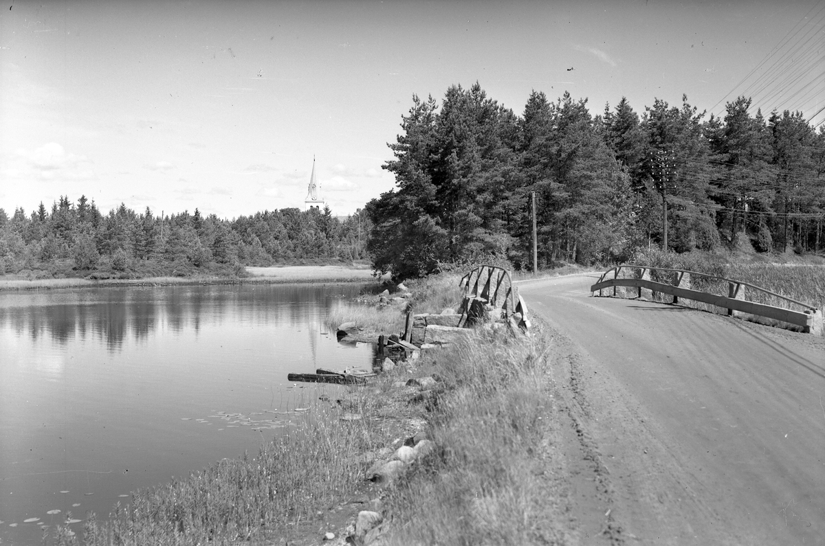 Vy över en sjö med en gammal bro till höger. I bakgrunden syns kyrktornet till Hinneryds kyrka.