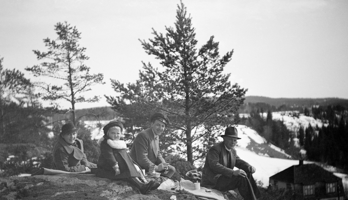 Bildet viser ei gruppe på fire personer på utflukt til en skråning like ovenfor et hus. Det er sannsynligvis på våren, for de sitter på bart berg, mens det fortsatt er snø på jordene i bakgrunnen. De har med utstyr til å drikke kaffe. Gruppa består av (fra venste): en kvinne med kåpe og hatt, ei jente med støvler, kåpe og hatt, en mann med bukse, jakke og sjåførlue, en mann med langbukse, jakke og hatt. Mannen med lua er Birger Christian Carlsen. I hånda holder han ei kameraveske. Fotografen er den femte i gruppa.