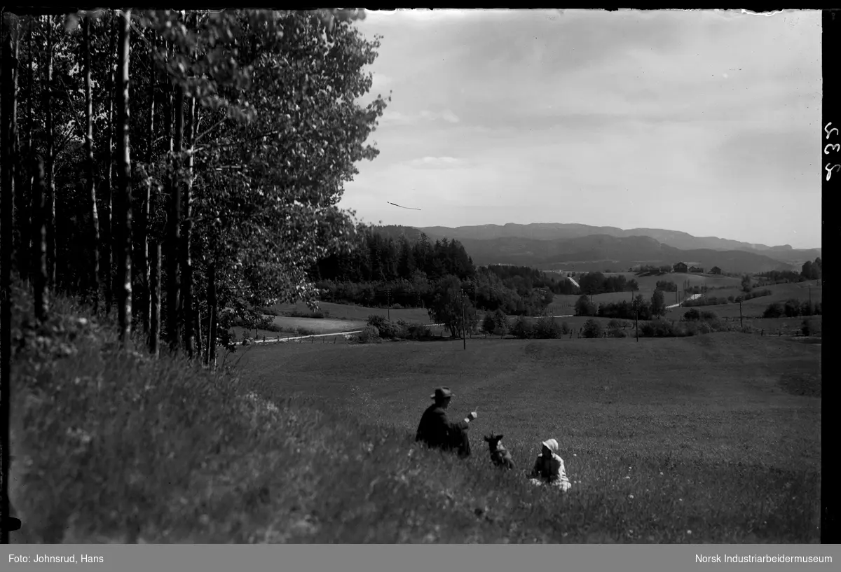 Utsikt mot Tinnegrend og Ramberg. Mann og dame med hund sitter i gresset.