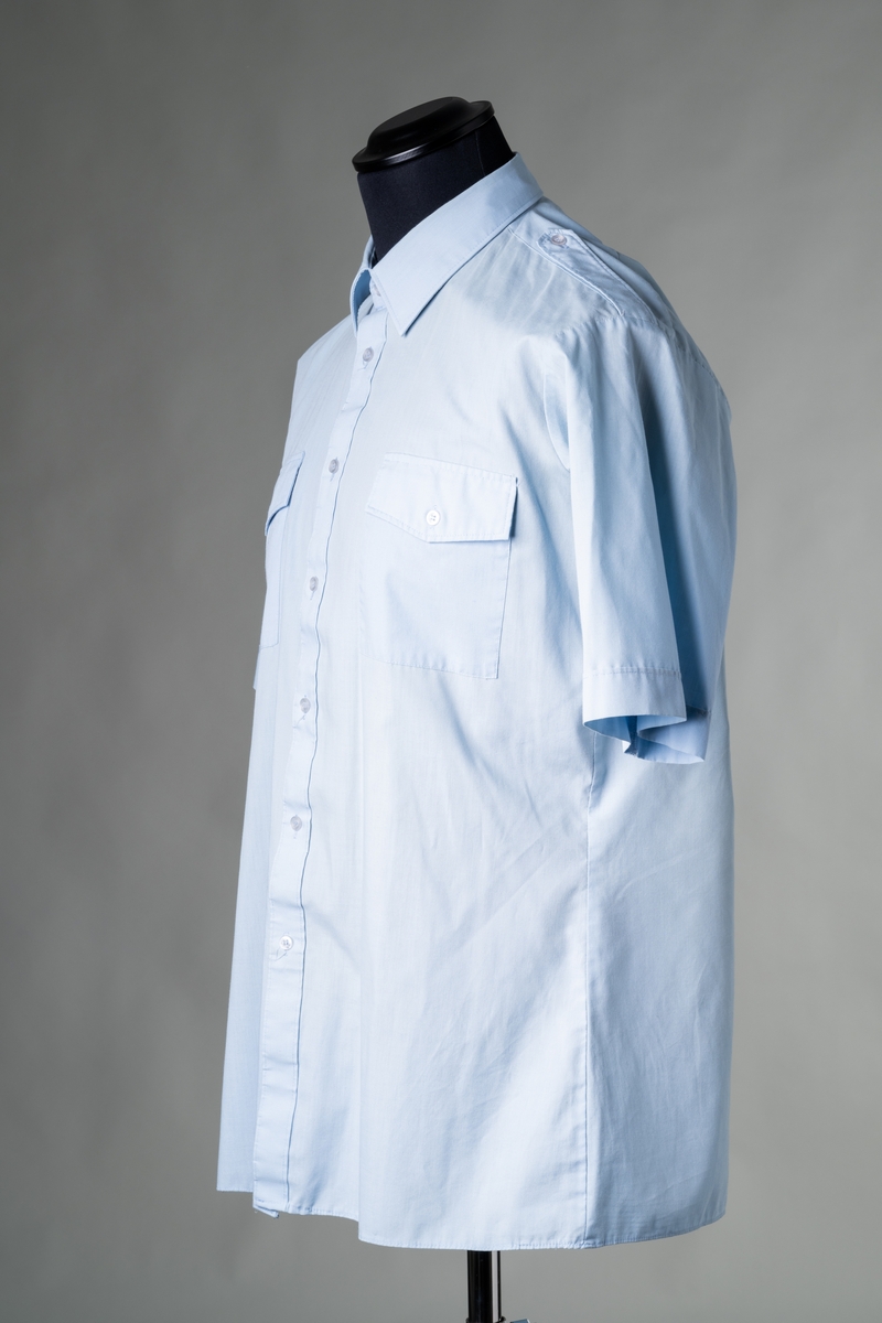 Skjorta till stationsuniform, modell sommar, för personal vid Brandförsvaret, Jönköpings kommun. Kortärmad skjorta av ljusblått tyg av polyester och bomull, insvängd midja, fast krage, ok av dubbelt tyg på ryggen. Knäppning fram med sju vita plastknappar, axelklaffar med en vit plastknapp, och två bröstfickor med lock och vit plastknapp. Tygetikett i nacken: "Bagutta". Storlek: C-bål, L, 42-42.

Samhör med: JM 56718:1-6.

Se vidare Historik.