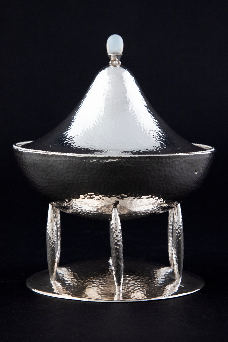 Bonbonnière med lokk i hamret sølv. Skålen er halvkuleformet og står på seks føtter festet til en sirkulær, svakt hevet fot. Lokket har konisk form og ender i en lokknapp med en opal. Langs kanten er lokket dekorert med perlestav.