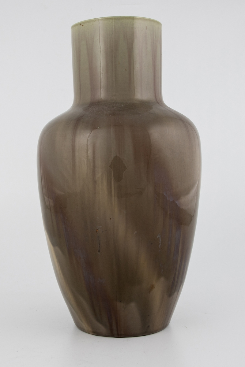 Vase i glasert flintgods. Urneformet koprus med sylindrisk hals. Vasen er dekket med grågrønn glasur, hvorpå rødbrune partier i form av rennende striper tidvis kommer til syne.