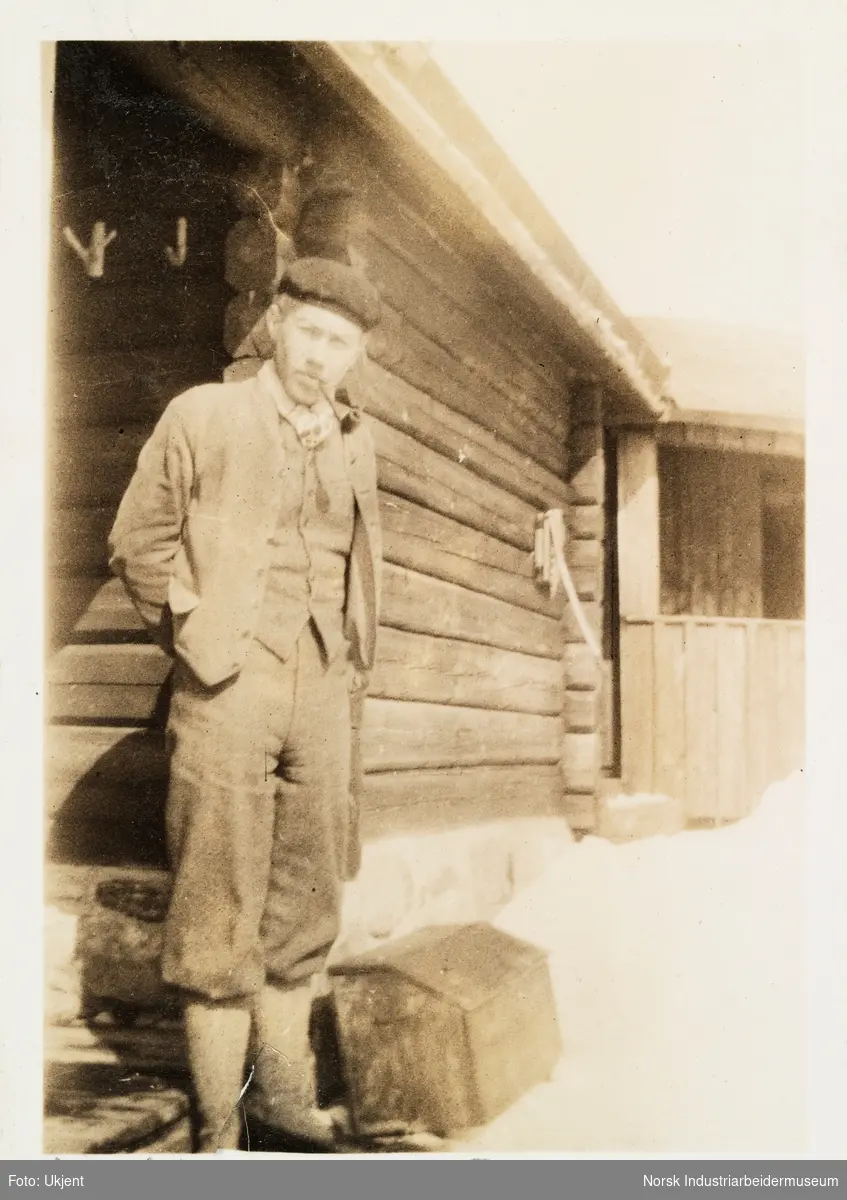 Påsken 1927 på Møsstrond. Mann i turtøy, sikspens og pipe står i snøen ved hjørne på laftet hytte på Møsstrond.