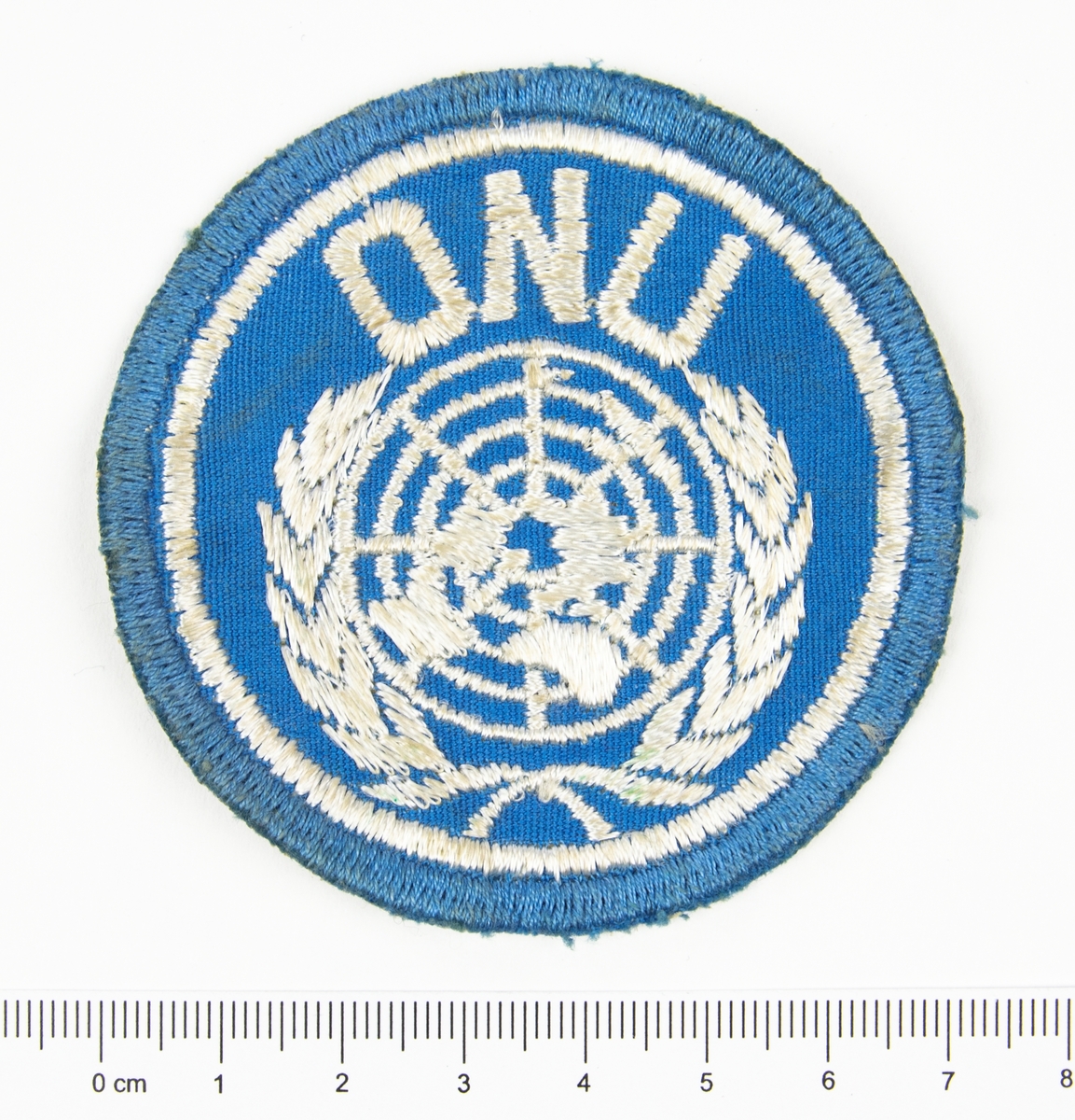 Förbandstecken. Textilt märke, ljusblått, med broderat vitt FN-emblem samt texten ONU.