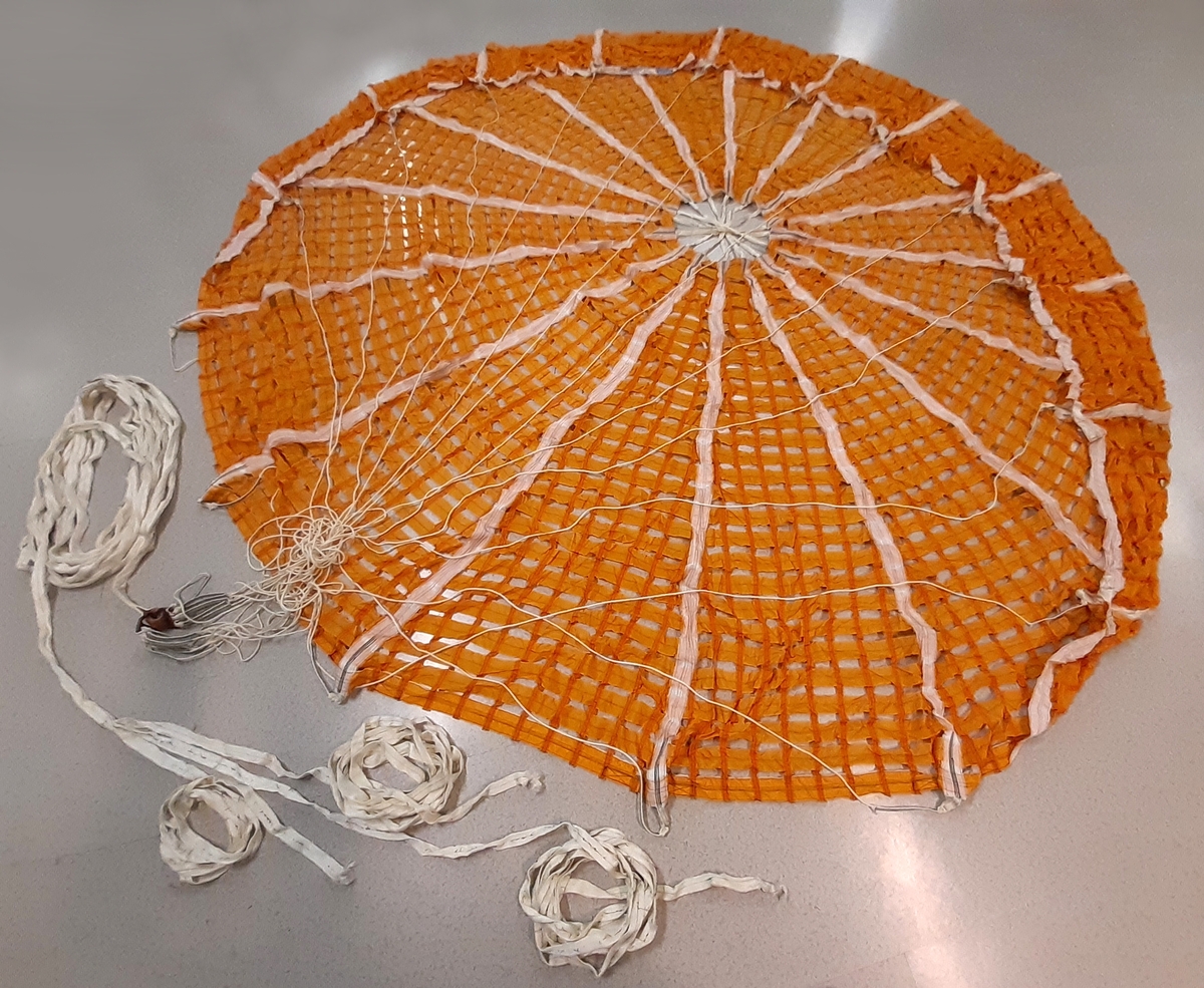 Antispinnskärm, bestående av orange textil samt vita linor. Medföljer en mindre vit skärm, märkt "IRVIN 2", denna sammanknuten med en brun textil påse. Samt en vit lina med stålvajer och metallfäste. Därtill två tygstycken med ögglor och ett mindre tillhörande textilt fragment.