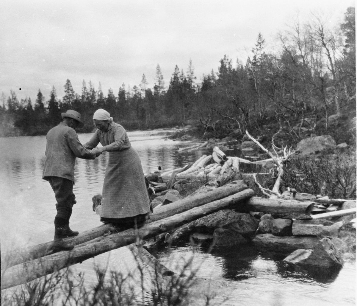 En mann og ei dame balanserer på tømmerstokker over ei elv