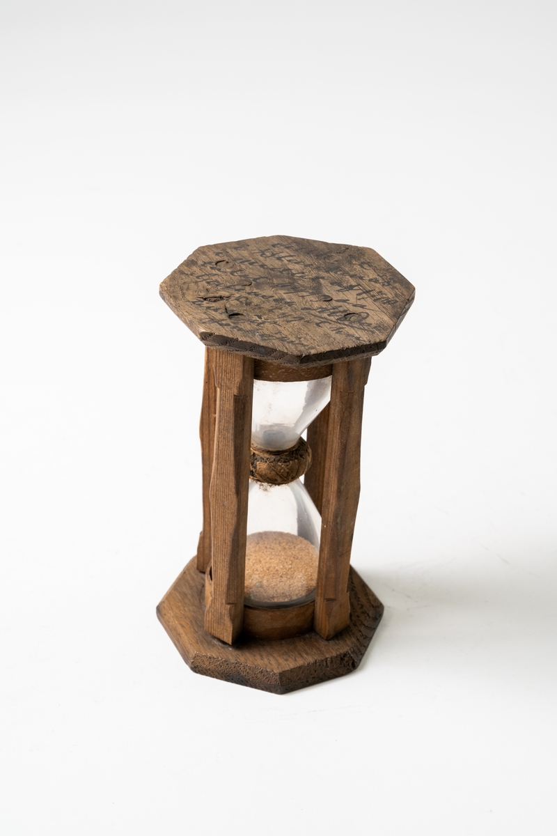 Timglas av glas, med sand som mätare, sitter i åttkantig träform som hålls ihop med fyra träribbor. Glaset hålls på plats med näver. Text på timglasets ovansida.