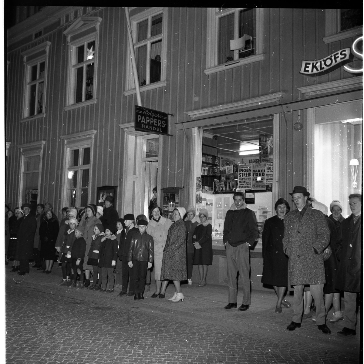 Grännabor, barn och vuxna, står utmed Brahegatan och väntar på Gränna scoutkårs lucia. Fasadskyltar för "Holgers Pappershandel" och "Eklöfs skor".