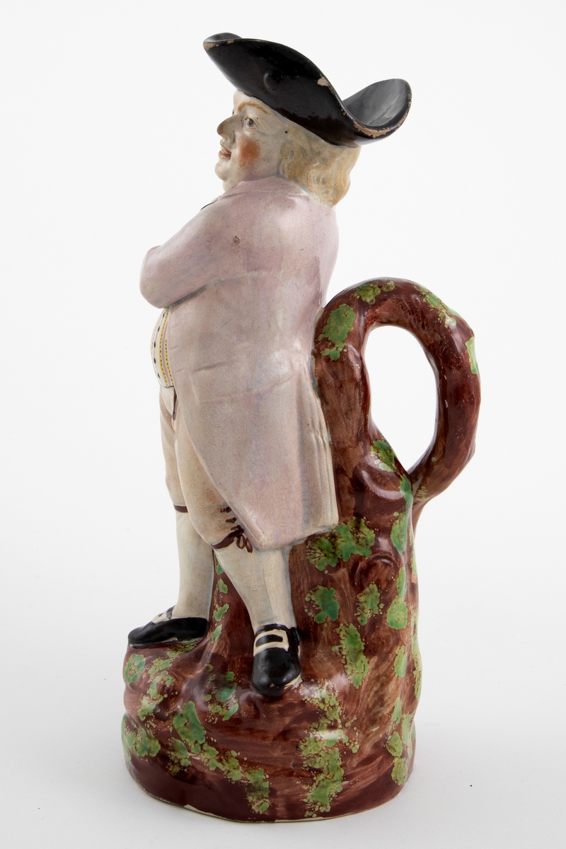 Toddykanne i form av en stående mannsfigur med pipe og kanne i hendene. Lokket mangler. Dekor i brunt, grønt, svart, gult og fiolett.