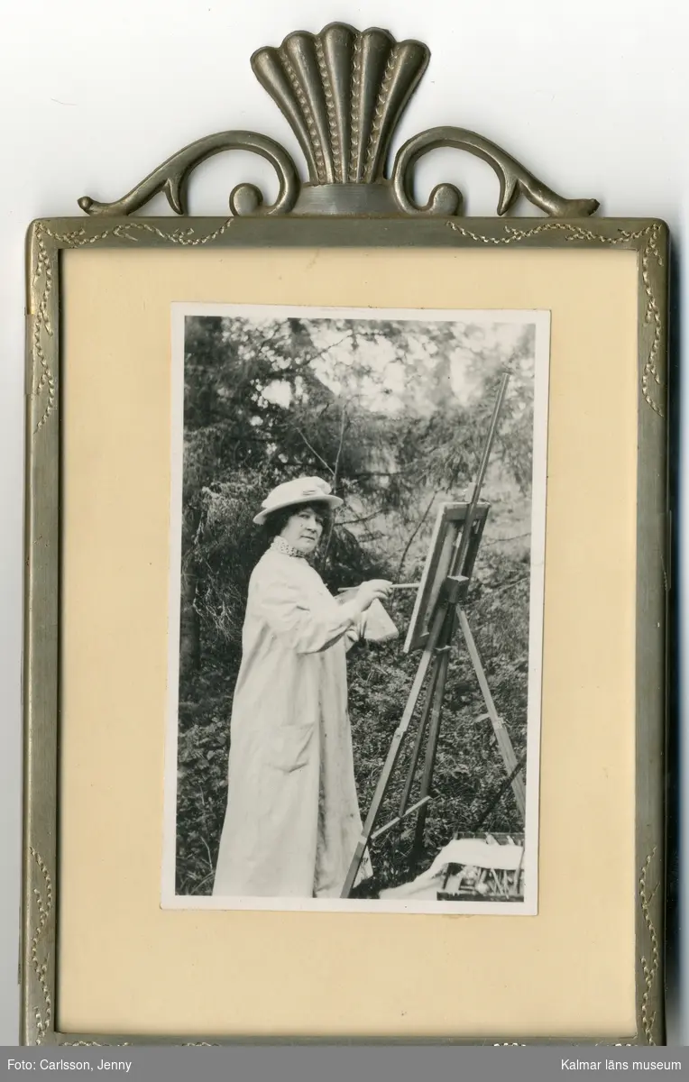 Jenny Nyström står vid ett staffli utomhus och målar. Hon är klädd i vit rock och vit hatt. Hon håller en palett i sin vänstra hand och en pensel i sin högra. Under staffliet ligger en låda med penslar m.m. på marken.