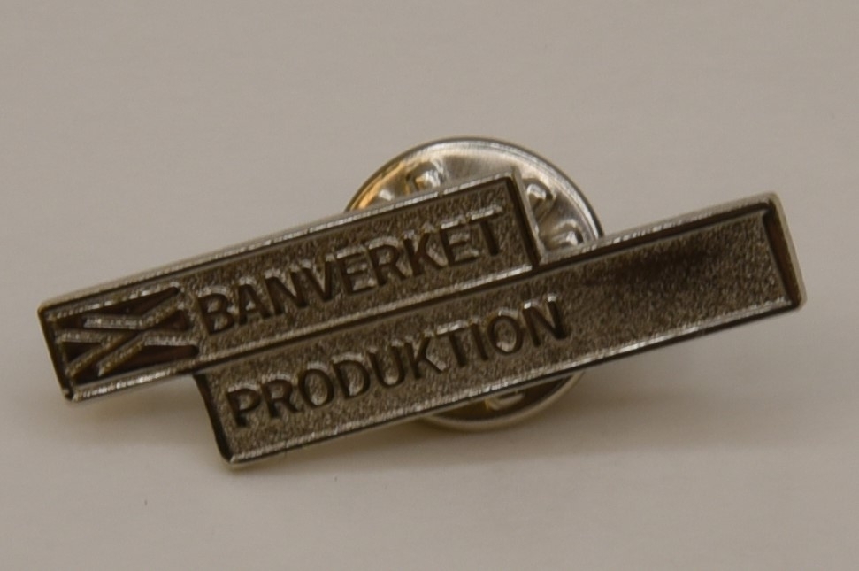 Pin av metall som består av två rektangulära rutor med text samt Banverkets logotyp i relief.(:1) På baksidan sitter en nål som fästs med hjälp av ett avtagbart fjärilslås.(:2)