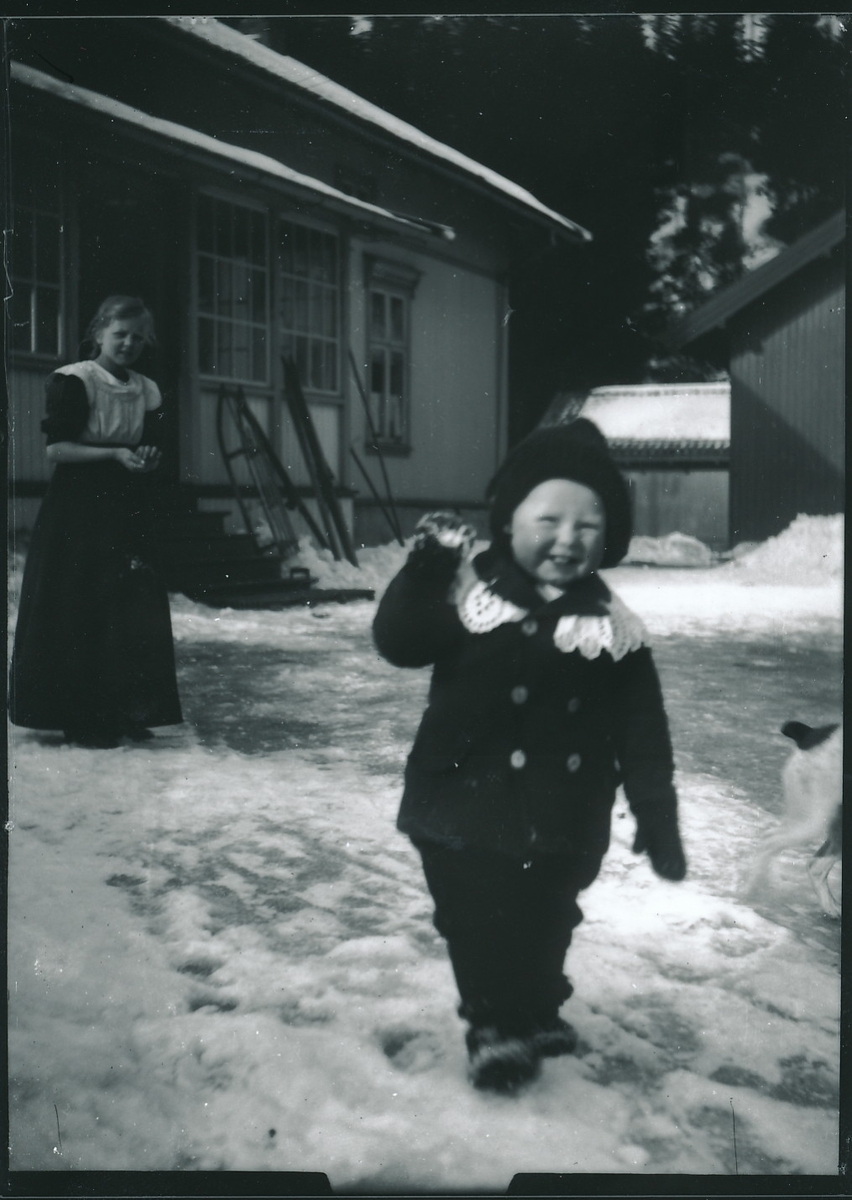 Wilhelm Fredrik Rydgren står i strikket antrekk på den snødekte gårdsplassen utenfor bestyrervillaen på Kistefos Træsliberi. Bak han står ei jente.  I bakgrunnen ligger bestyrervillaen med glassveranda og uthus. Ski og kjelke står lent mot husveggen.