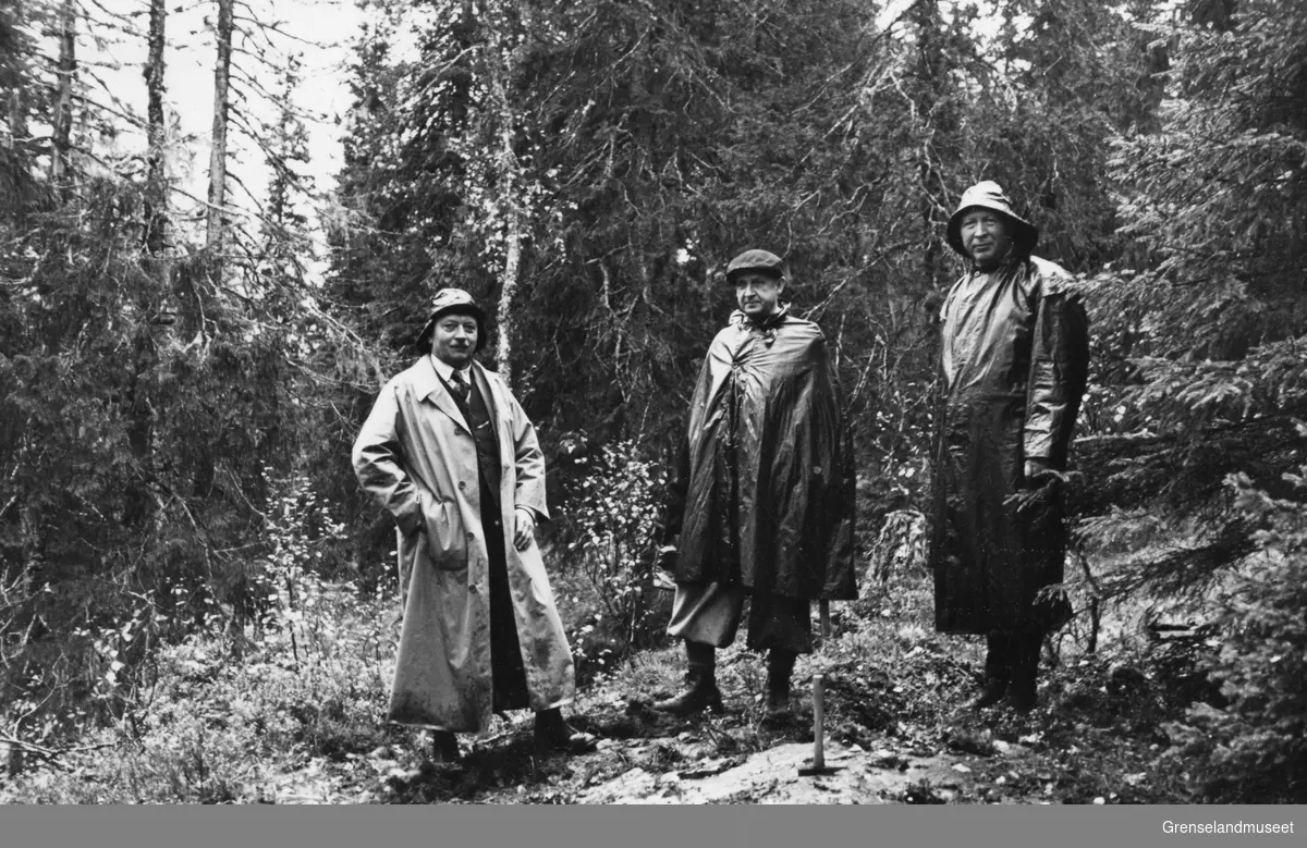 Tre menn står i en granskog foran et bart bergparti, en hakke tyder på at de tar mineralprøver, Rana september 1937.
