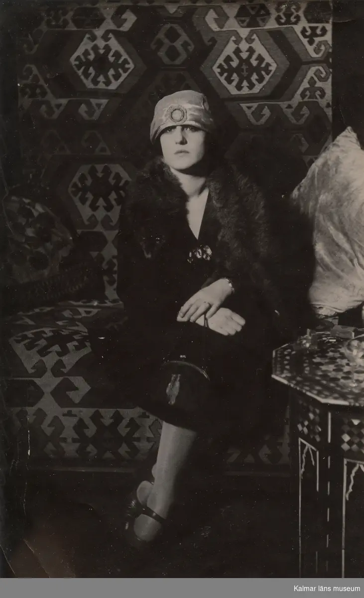 Fotografi av en ung kvinna klädd i 1920-tals kläder. Hon sitter på en soffa med ett bord till höger. [Ej signerad påskrift på baksidan av kortet: "Detta är taget på ateljén, rökbordet är något alldeles enastående vackert med pärlemor-inlägg, äkta."..