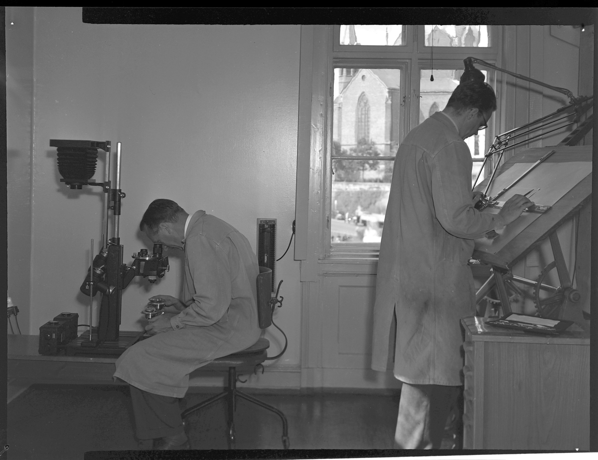 To mannlige laboranter i arbeid på Kriminallaboratoriet. Den ene kikker i et stort sammenligningsmikroskop. Den andre står ved et tegnebrett. Utsikt til Trefoldighetskirken gjennom vinduet.