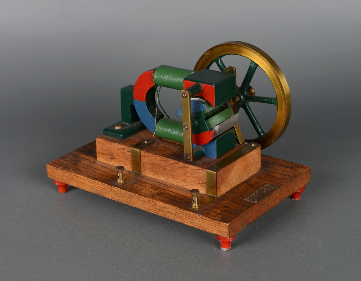 Et apparat til fysikkundervisning som viser hvordan elektromagnetisme fungerer. En treplate som står på fire foter med en trekloss limt og skrudd fast. På treklossen er apparatet av grønnmalt/lakkert jern med hjul, magneter og isolert kobbertråd skrudd fast.