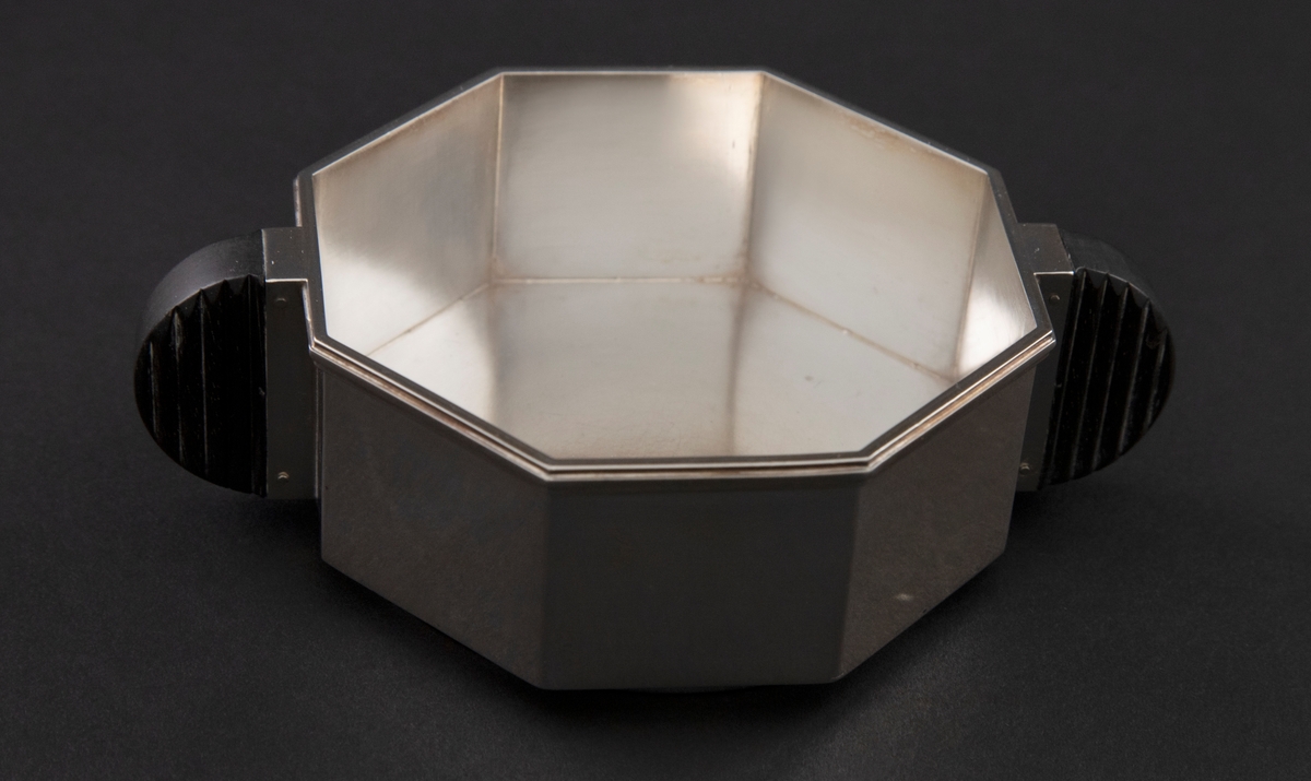 Liten sølvskål med oktogonal grunnform og rette sider. På korpus er festet to halvsirkelformede håndtak i mørkt tre.