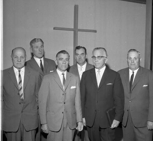 Sex herrar i mörka kostymer och slips står uppställda framför ett kors.