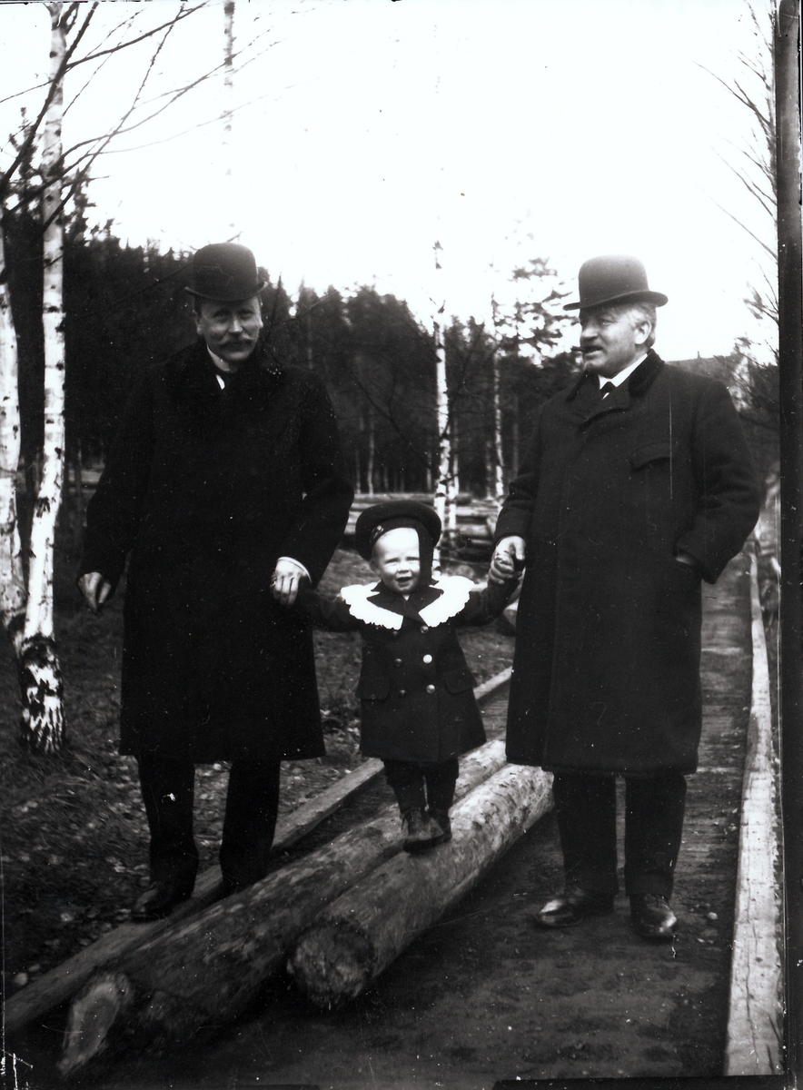 Bestyrer Wilhelm Rydgren (til høyre) sammen sin yngre bror Thorvald Rydgren på tur på området til A/S Kistefos Træsliberi med Wilhelm Fredrik mellom seg. De går på en gangsti med tømmerstokker til venstre. I bakgrunnen er lagring av tømmerstokker. Til høyre i bildet, over skulderen til bestyrer Rydgren, skimtes antagelig bestyrervillaen.
