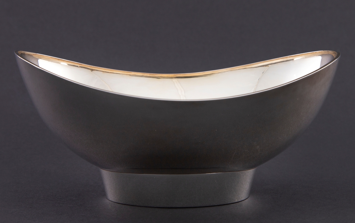 Skål i sølv med oval form. Den står på en oval fotring, samt har bølgende formet munningsrand. Innvendig er skålen forgylt.