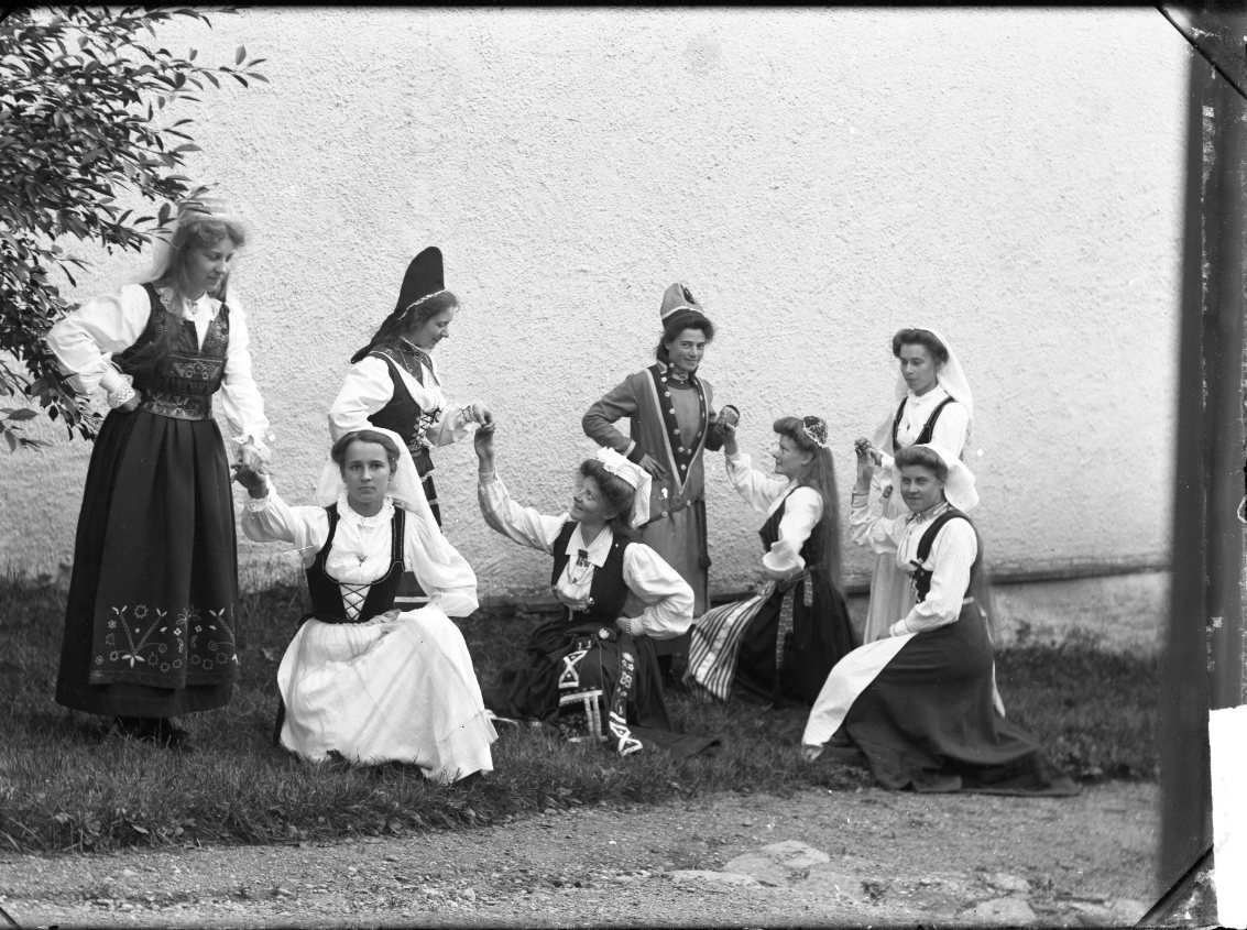 Åtta kvinnor i folkdräkter i uppvaktande poser: "Sällskapsteatern Grenna".