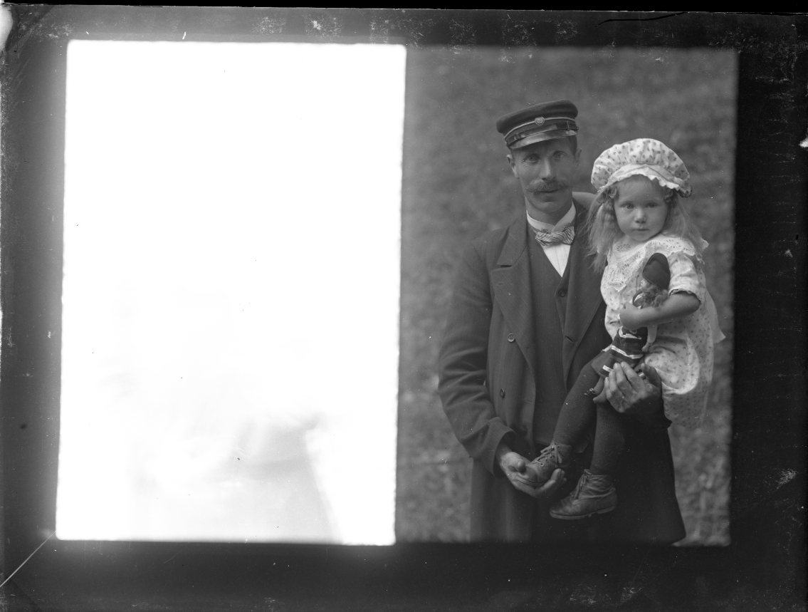 En man med mustasch och keps håller en flicka i famnen, hon i sin tur har en docka i famnen.