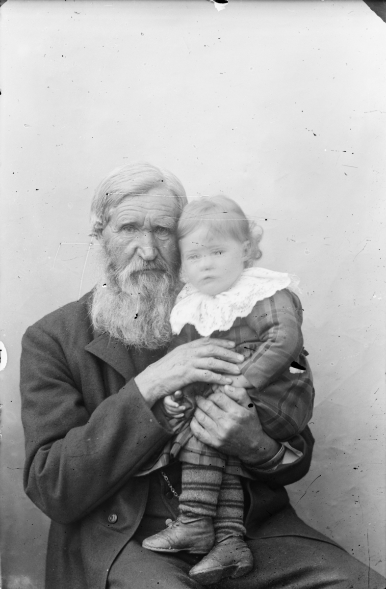 Eldre mann i halvfigur med  med et barn på armen. Barnet har kjole med stor bolondekrage og sko. Han har grått hår og stort sjegg, kledd i mørke bukse og vadmelsjakke