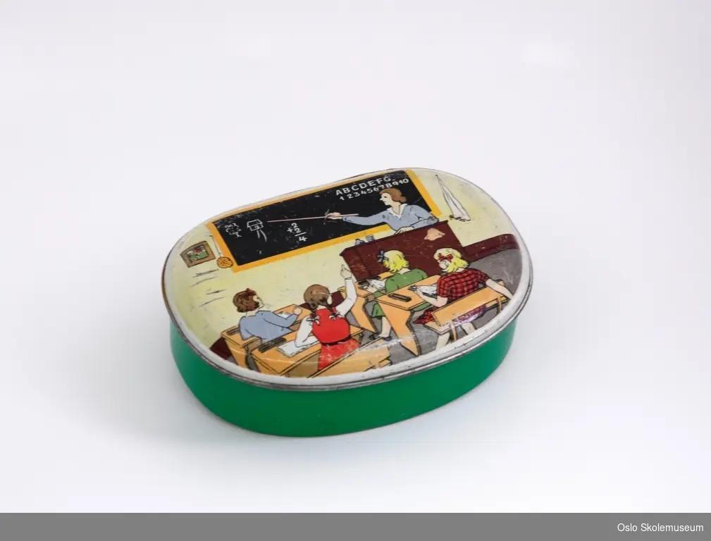 Oval matboks i blikk med lokk som er festet på en ene langsiden. Selve boksen er grønn. På lokket er et motiv av et klasserom med en lærer og fire elever.