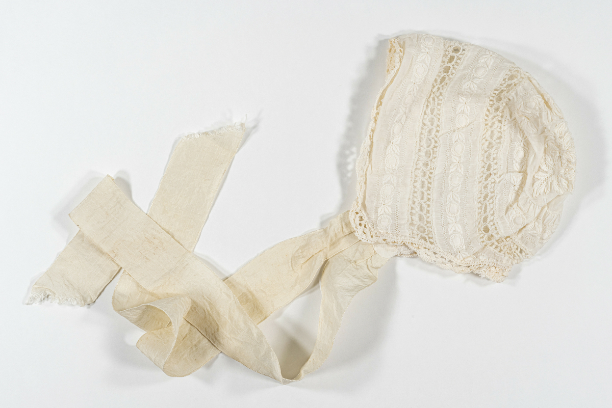 Lue (dåpslue?) i hvit bomull/blonde (?) med heklede partier. Påsydd snørebånd rundt halskant i silke. 
Har tilhørt Sara Fabricius.