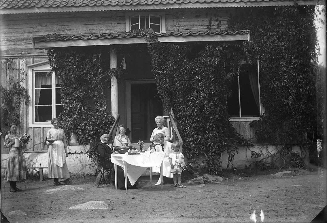 Fyra kvinnor och ett barn sitter runt ett kaffebord på en gårdsplan framför en farstu. En äldre kvinna är klädd i mörkt, troligen "friherrinnnan Falkenberg" från Stockholm.
Till vänster, en liten bit från sällskapet, står två flickor med en karaff och blommor i vas respektive bricka